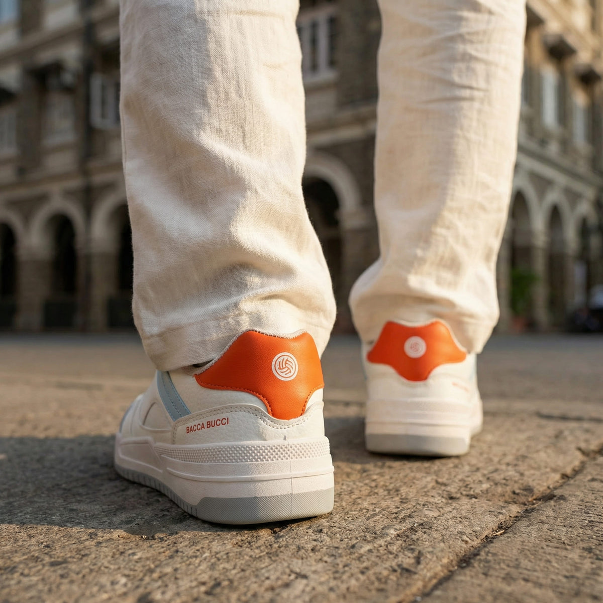 Bacca Bucci white sneakers with orange accents worn on a street with a blurred building background.